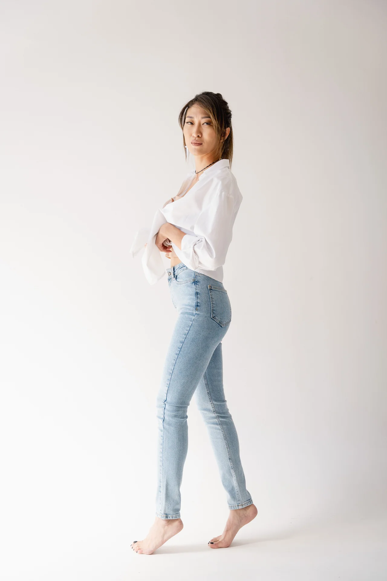 Woman wearing light blue slim-leg jeans and a white blouse against a pale studio background.