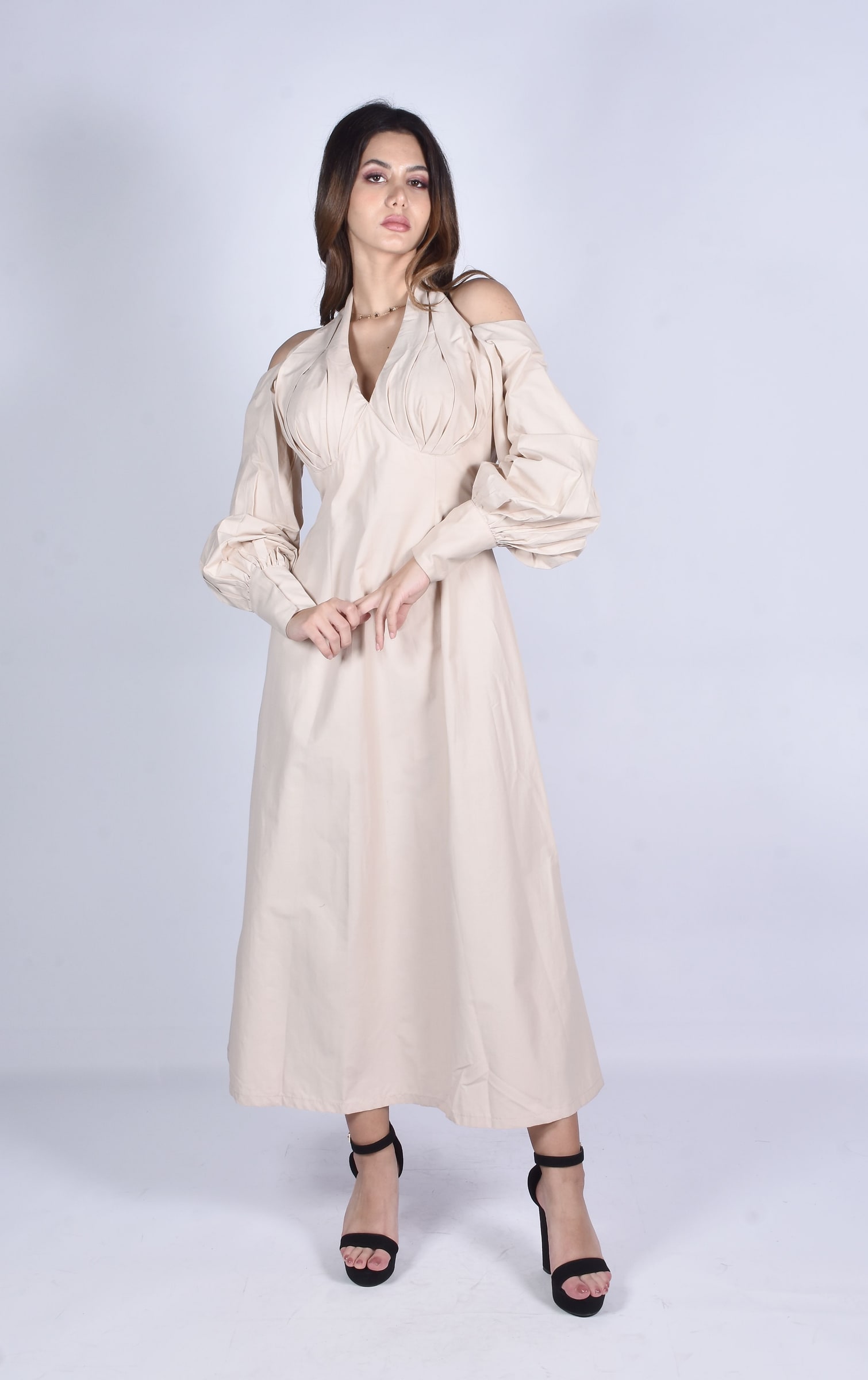 Woman wearing a light beige long-sleeve midi dress with open shoulder details against a pale studio background.