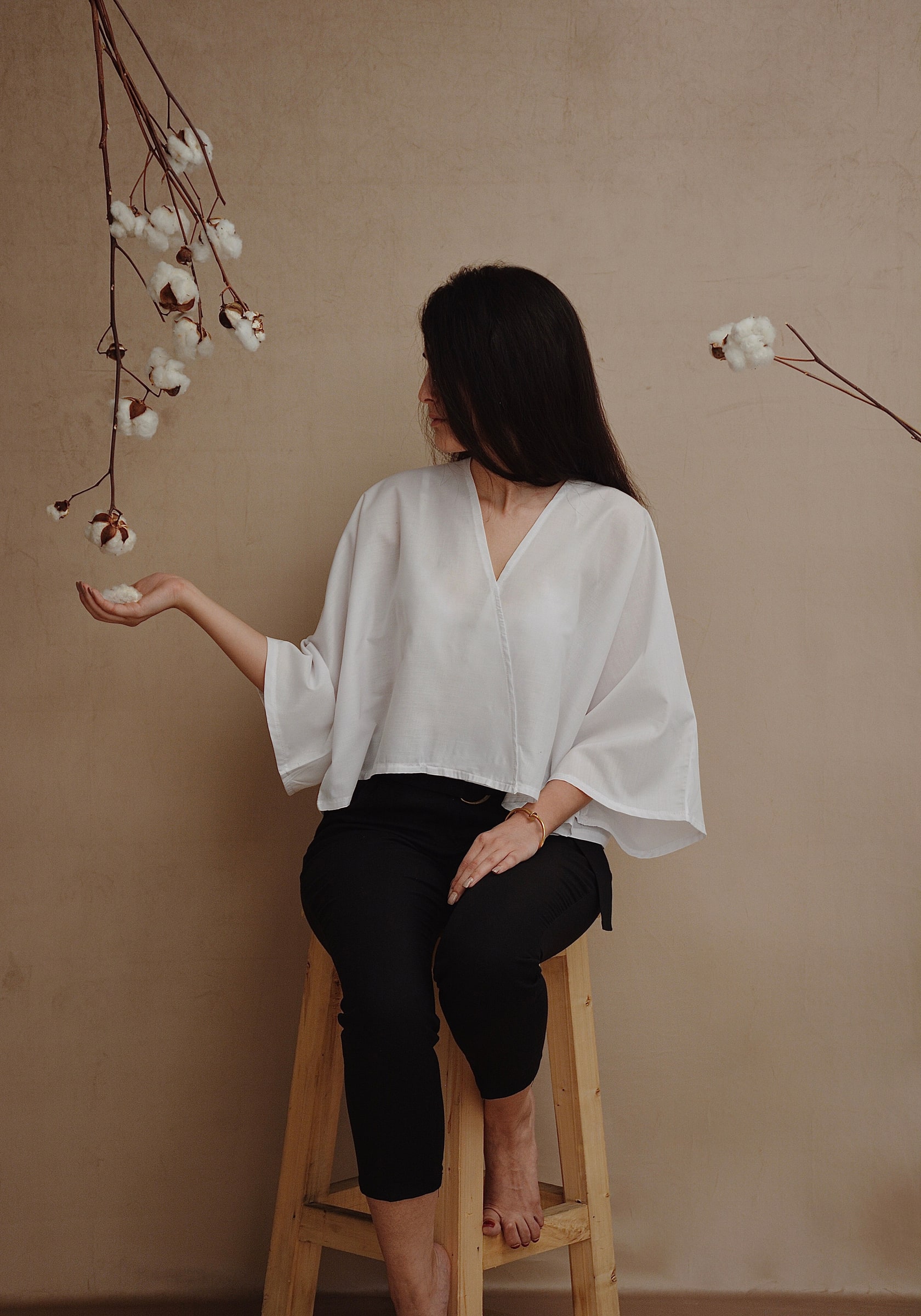 Woman in a loose white V-neck blouse with wide sleeves, seated against a warm neutral studio background.