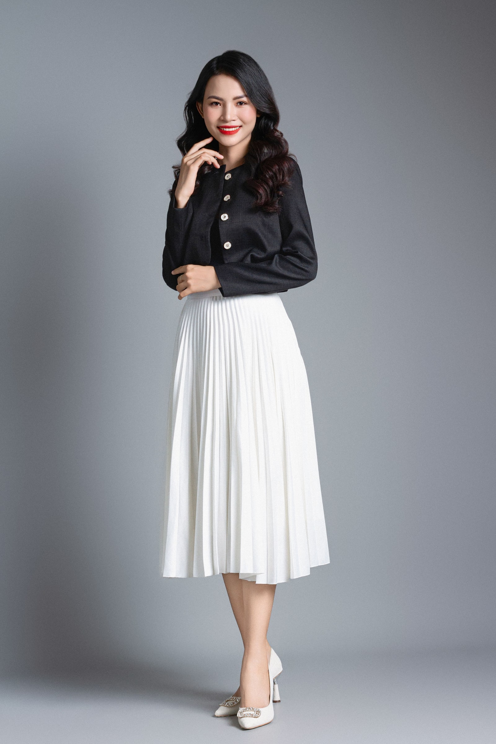 Woman wearing a white pleated midi skirt and dark cropped jacket against a neutral grey studio background.