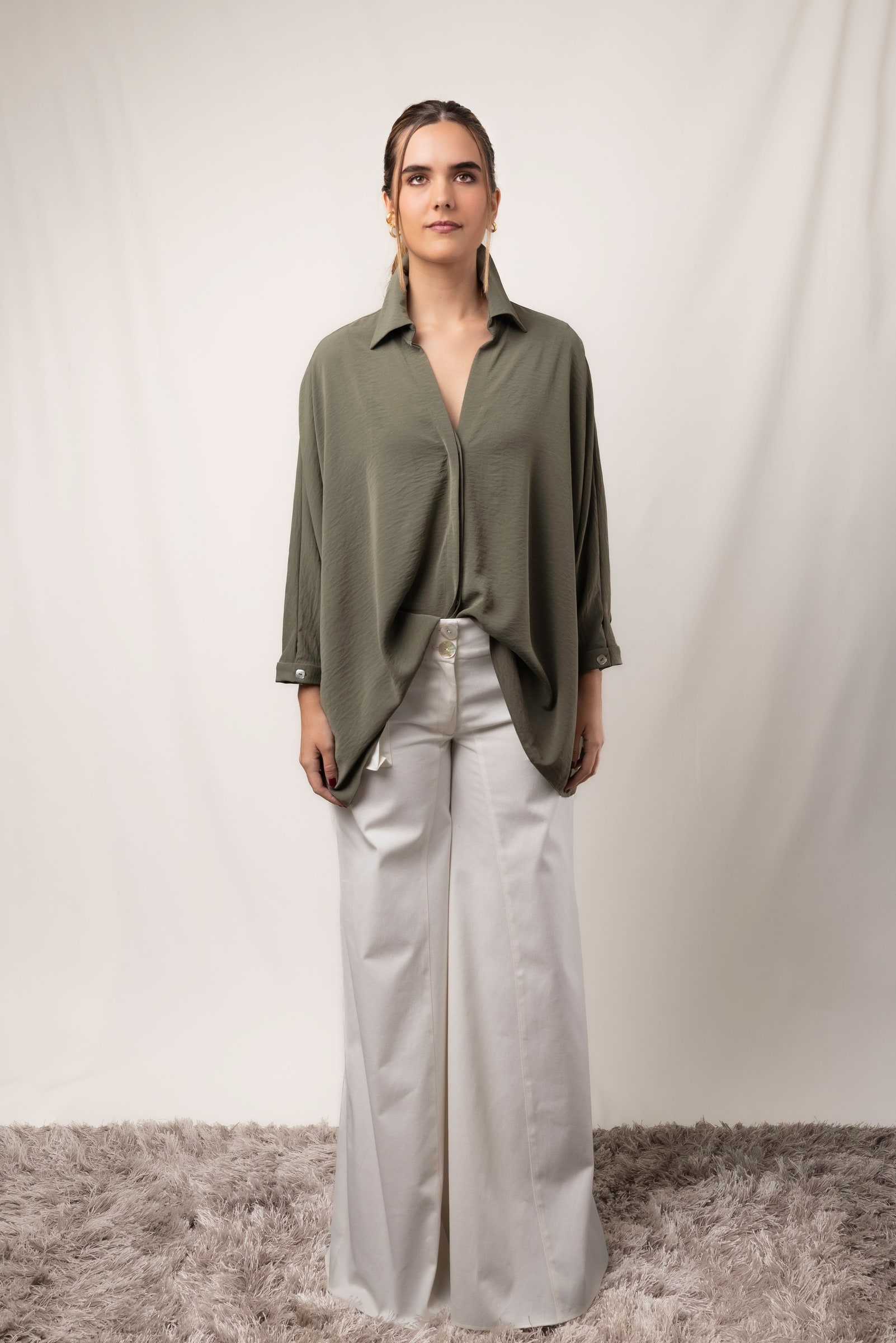 Woman wearing light grey wide-leg trousers and a relaxed olive shirt against a warm neutral studio background.