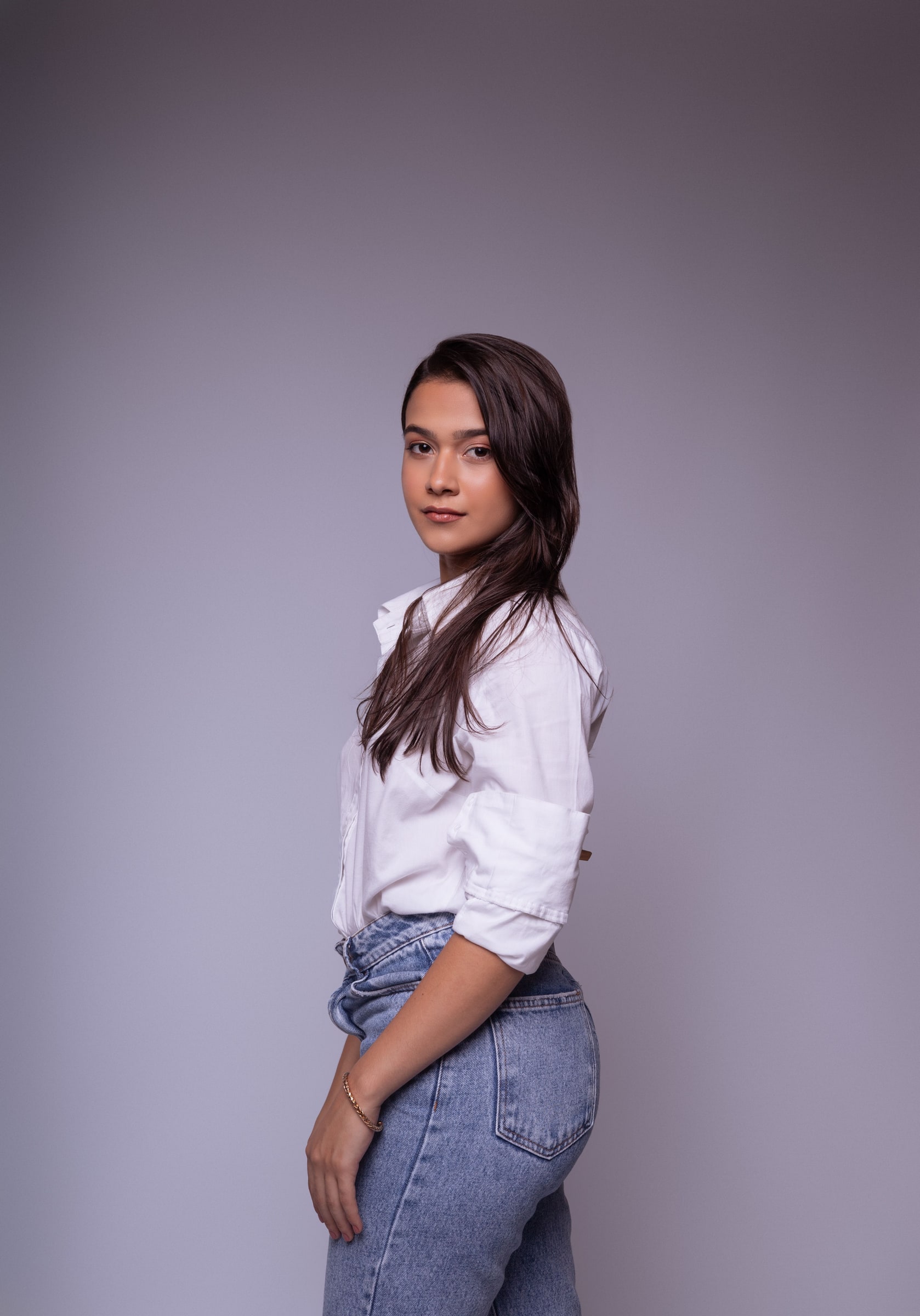 Woman wearing a light button-front shirt with rolled sleeves and jeans against a soft grey studio background.