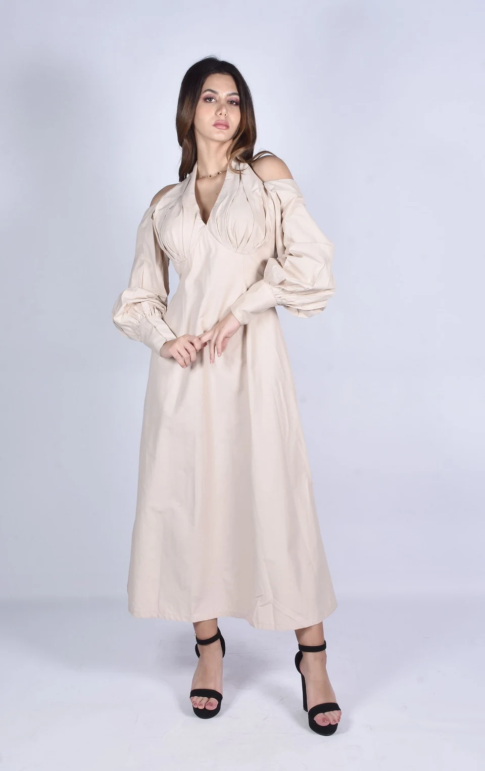 Woman wearing a light beige long-sleeve midi dress with open shoulder details against a pale studio background.