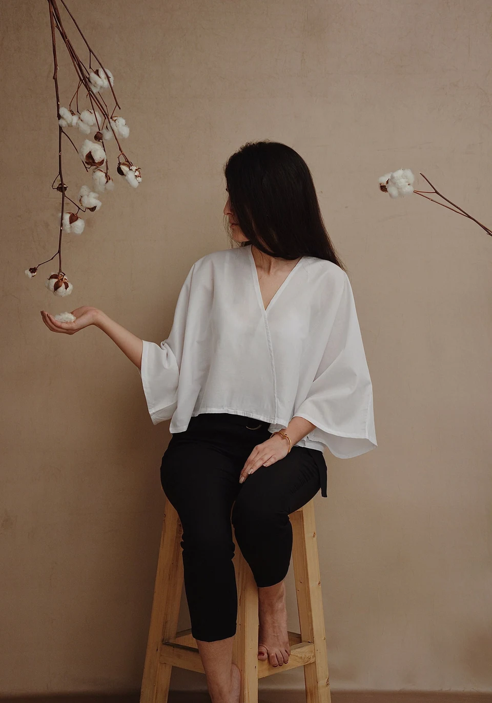 Woman in a loose white V-neck blouse with wide sleeves, seated against a warm neutral studio background.