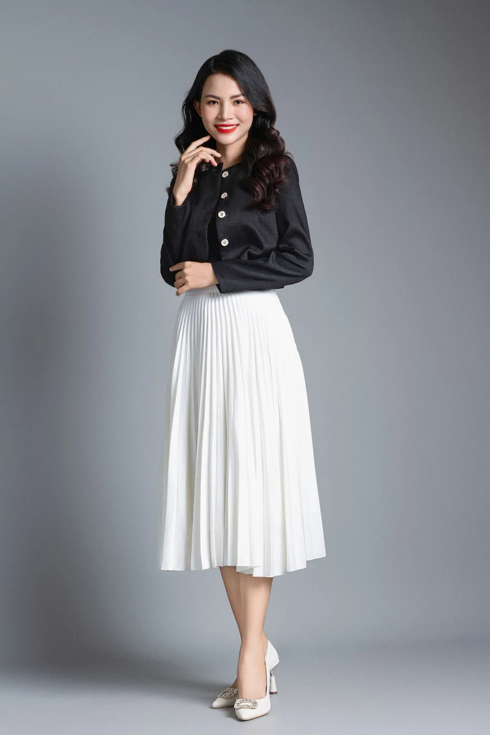 Woman wearing a white pleated midi skirt and dark cropped jacket against a neutral grey studio background.